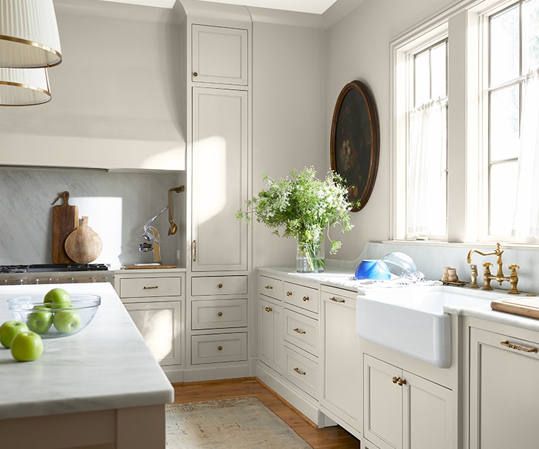 White-painted kitchen with ample counter space, farmhouse sink, kitchen island, and various plants and flowers.