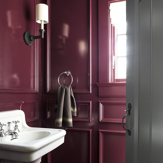 A bathroom with plum-painted panelled walls and trim, a white pedestal sink and gray-painted door.