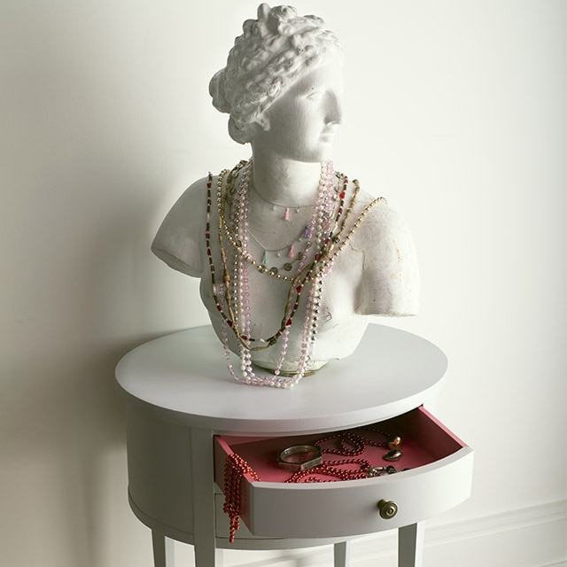 Un buste sculpté paré de bijoux sur une table ronde peinte en gris clair avec un tiroir ouvert, devant un mur peint en blanc.