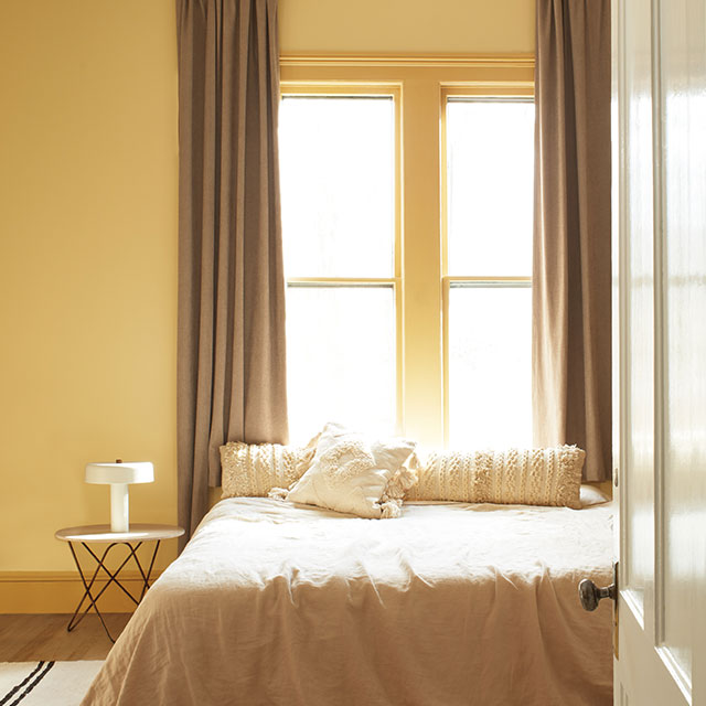 A gold-painted bedroom with a bed on a striped rug in front of a large window with beige curtains, featuring an end table and lamp.