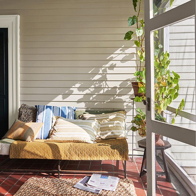 An outdoor room with white siding, plants, a couch with mismatched pillows and yellow blanket, and a rug.