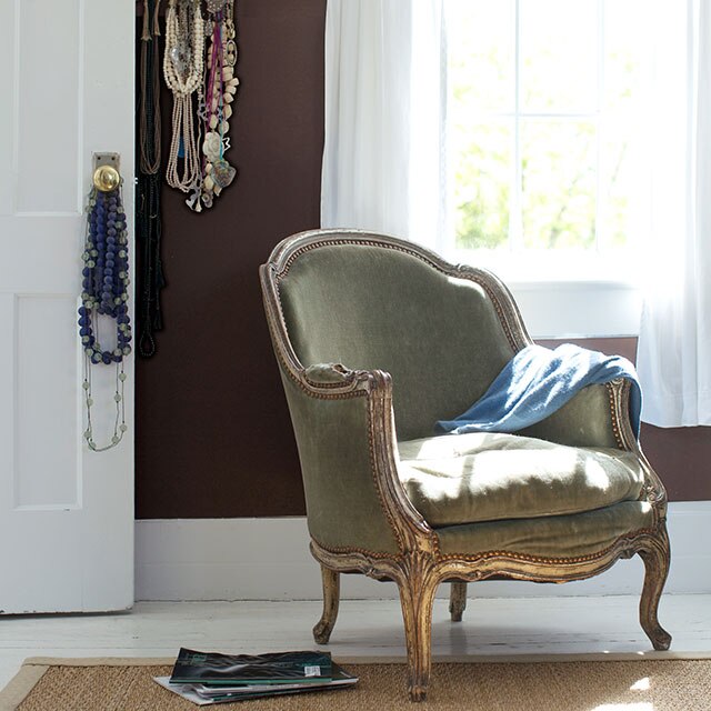 A gray vintage-style chair in a brown-painted room with various necklaces on the wall and hung on a doorknob on a white door.