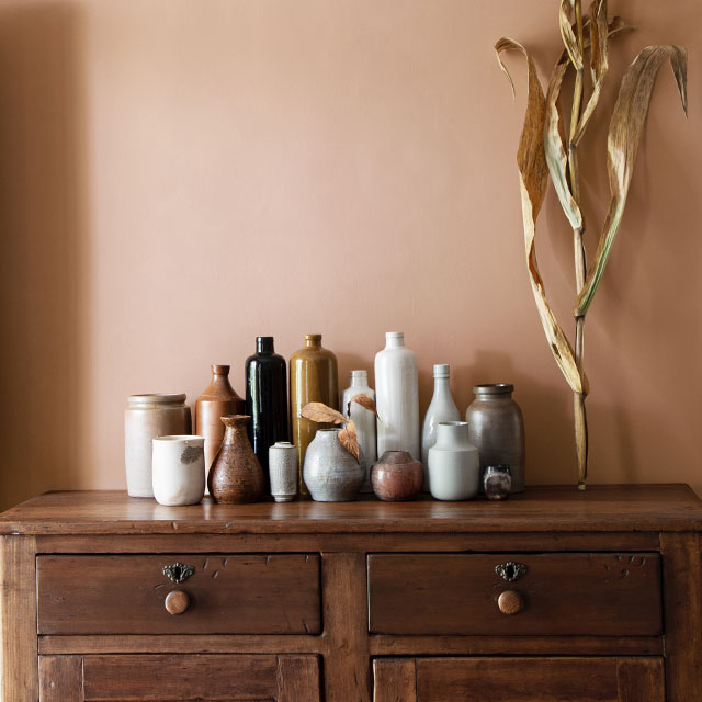  Un vase, une cruche et un bol posés sur un buffet en bois contre un mur brun pâle nuancé de rose.