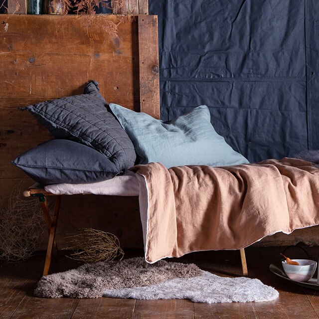A wooden chaise with linen bedding and blue fabric background.