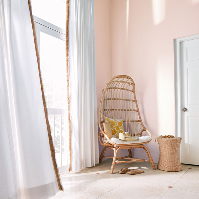 Murs peints en rose, plafond et moulures blancs, fauteuil en osier avec coussin jaune et rideaux vaporeux blancs.