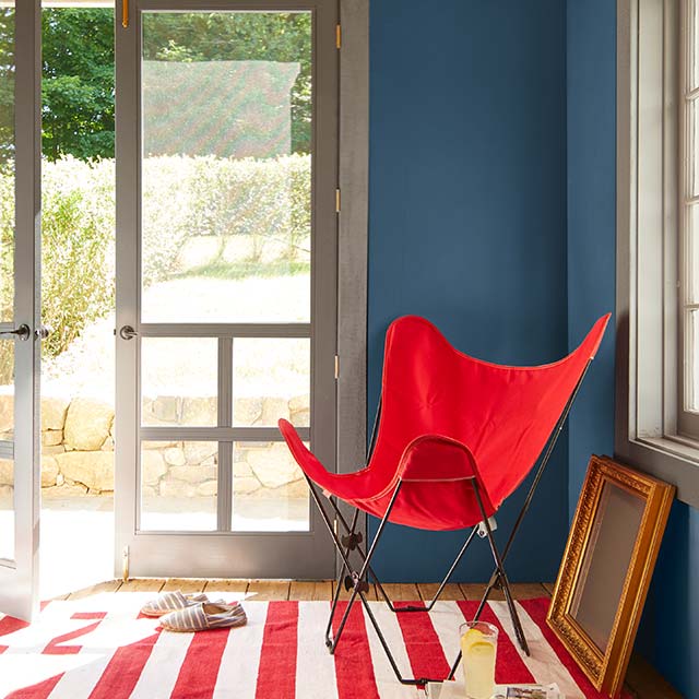 Une entrée aux murs peints en bleu avec une porte moustiquaire à deux vantaux aux cadres blancs, un petit tapis rayé et un fauteuil en toile rouge.