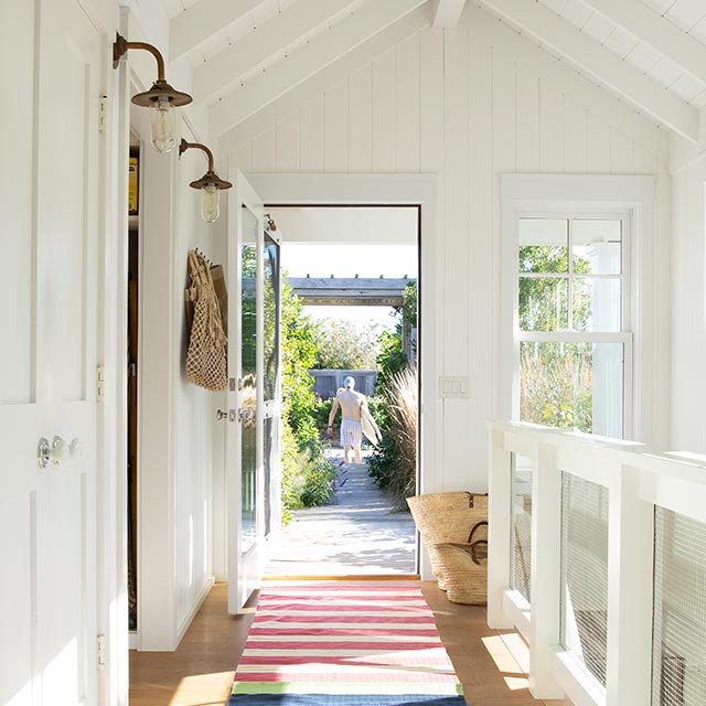 L’entrée de cette maison en bord de mer a des murs blancs, une balustrade en câble métallique, un passage à rayures et un plancher en bois clair.