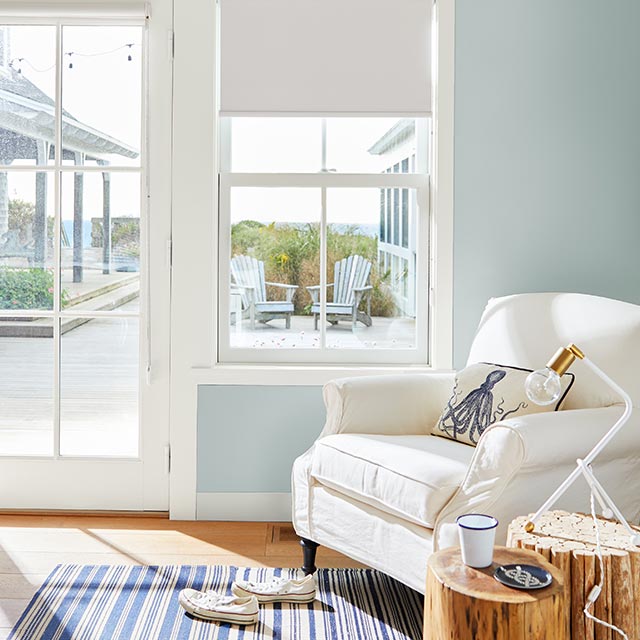 Salon ensoleillé peint en bleu pâle avec fauteuil blanc rembourré et vue sur la mer à travers une porte vitrée et des fenêtres.