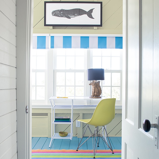 Una puerta y una pared de pasillo pintadas de blanco abren paso a una oficina en casa de ambiente costero con una pared y un techo de traslapados pintados de amarillo pálido, un cuadro de una ballena que cuelga sobre las ventanas, un escritorio blanco con una silla amarilla y un piso pintado de azul.