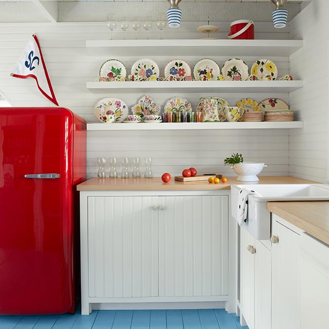 A white-painted kitchen with light blue-painted floors, red refrigerator and floating shelves.