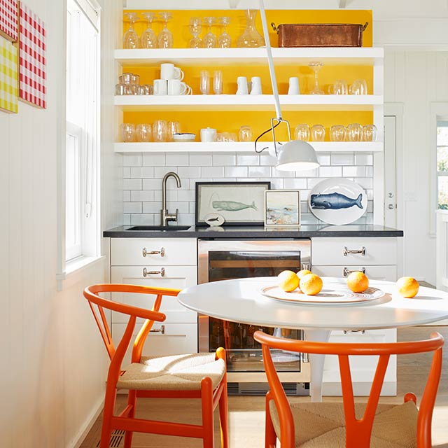 A small, white-painted kitchen with yellow shelving accent wall and orange chairs around a white tulip table.