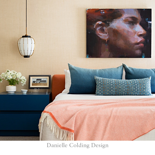 Bedroom with apricot throw, orange bed base, blue accent pillows and wall art of a woman; pendant light over blue side table.