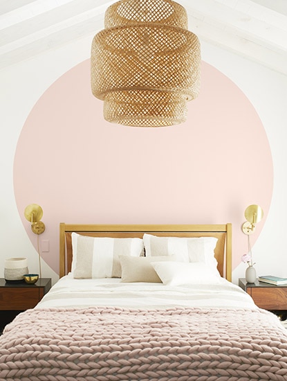 Bedroom with white walls and a large light pink circle behind white bed with pink throw blanket and a basket chandelier.