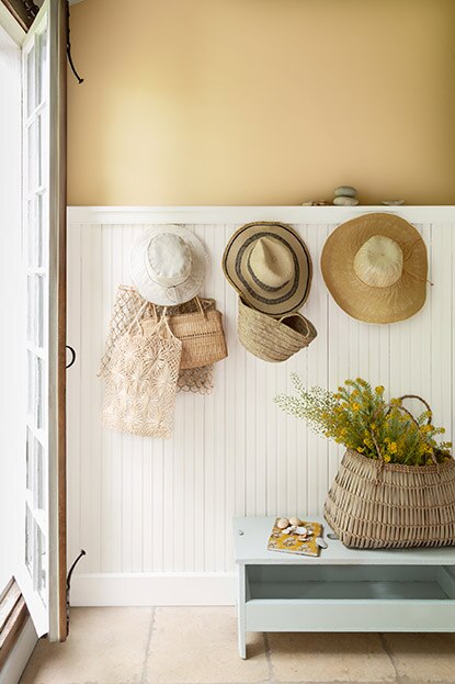 Trois chapeaux et des sacs sont accrochés sur les lambris de bois blancs recouvrant les trois quarts de la hauteur d’un mur beige doré.