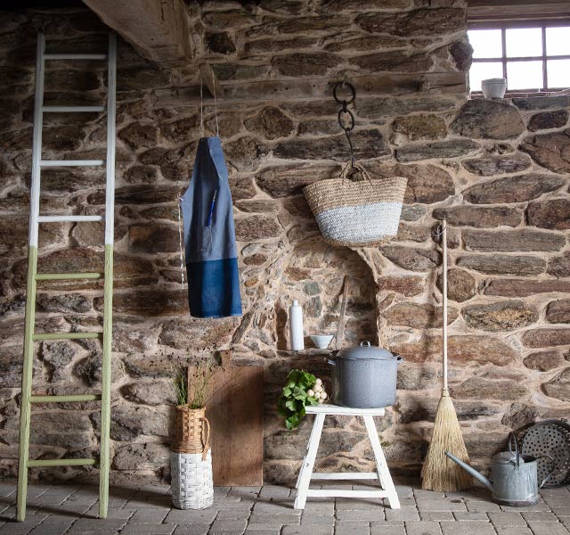Ladder leaning on a stone wall next to a broom, wicker basket, watering can, and stool topped with flowers and a pot.