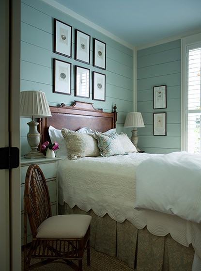 Blue/green bedroom walls with six picture frames hung above a cottage inspired bed with white bedding.