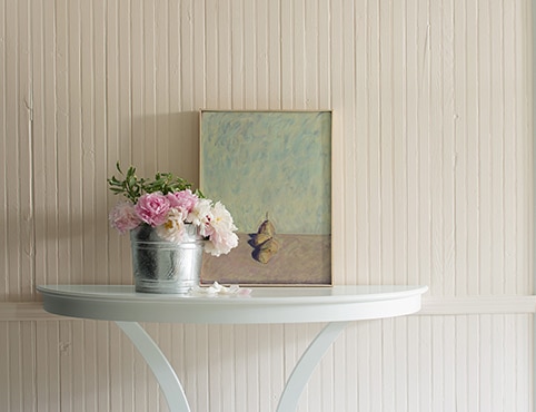 Pale pink walls with a painting and a tin bucket of pink flowers on top of a console.