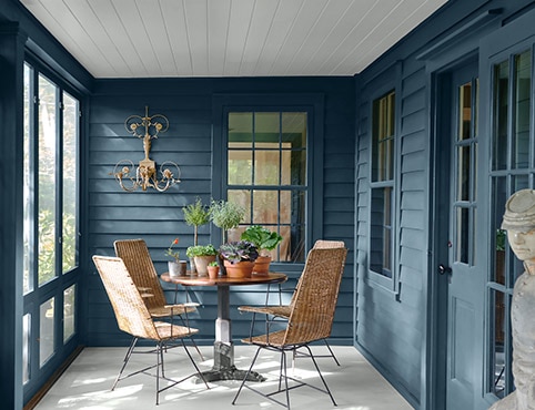 Le porche fermé d’un chalet peint en bleu, une table moderne, des chaises, des plantes d’intérieur et une applique murale.