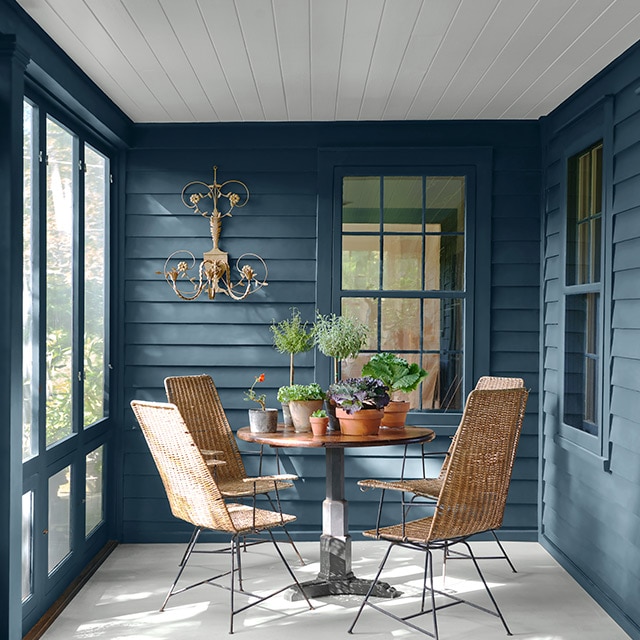 Le porche fermé d’un chalet peint en bleu, une table moderne, des chaises, des plantes d’intérieur et une applique murale.