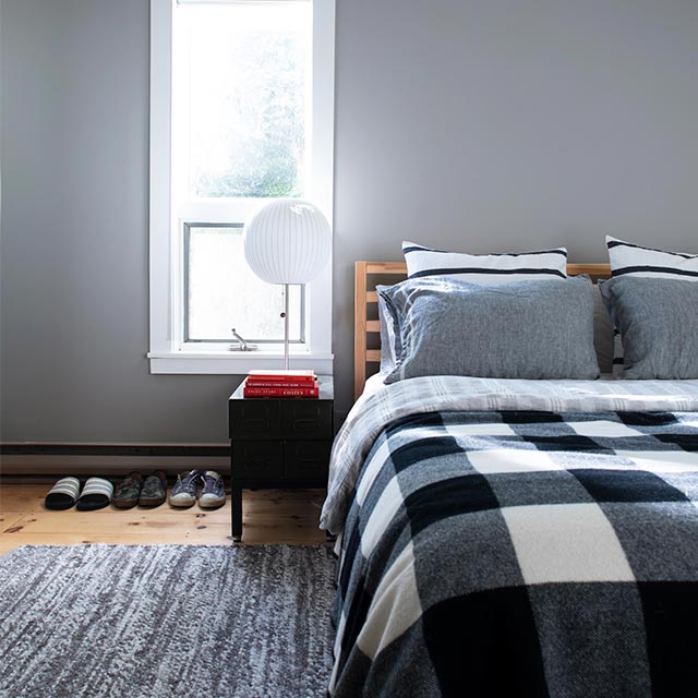 Gray cabin bedroom with white trim and shiplap, stained wooden beams, plaid bedding, sphere lamp, and shoes.