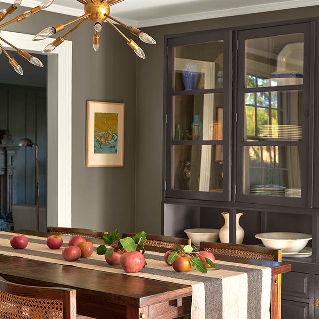 Un vaisselier brun dans une salle à manger peinte en gris  arborant une table en bois et un chemin de table rayé, des luminaires éclectiques et des moulures peintes en blanc.