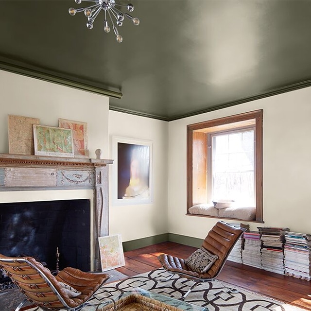 A cozy, sunlit living room with white-painted walls, a dark green ceiling and trim, leather chairs and printed rug, a wood mantel, window trim and door.