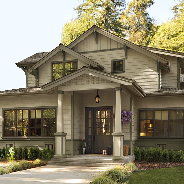 A beautiful taupe-painted Craftsman style home with gabled rooflines and a front porch, a black front door and sage accents and trim, surrounded by greenery.