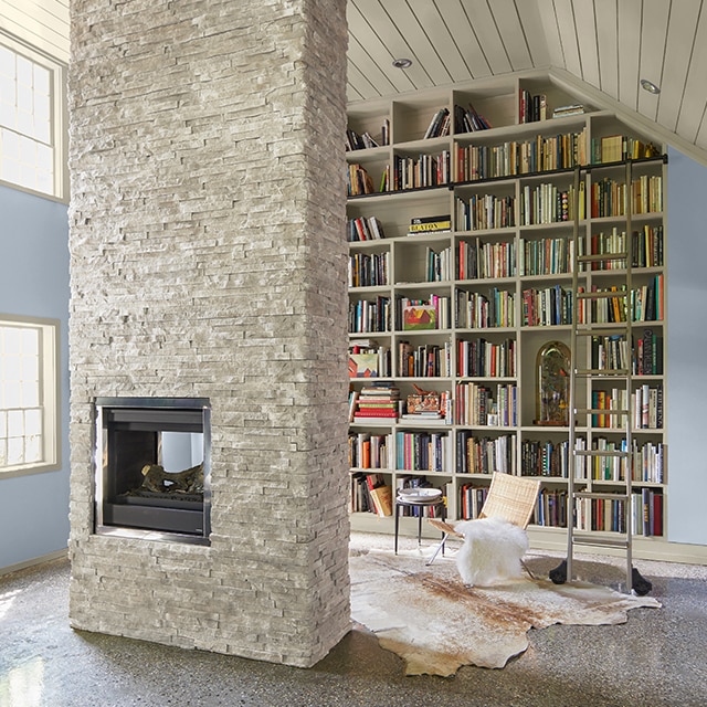 A home library with a statement floor-to-ceiling white-painted bookcase, blue walls, white shiplap vaulted ceiling and trim, and white stone center fireplace.