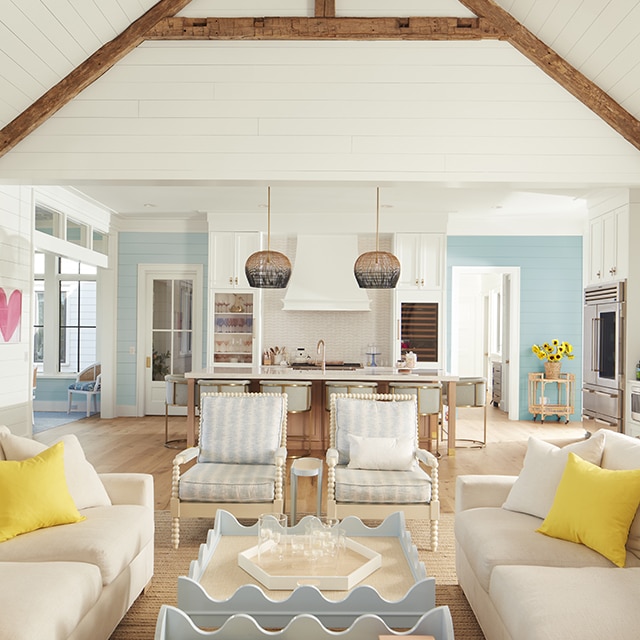 An airy, open white-painted living room and kitchen with shiplap walls and vaulted ceiling, wood beams, blue and white decor, and far back blue walls.