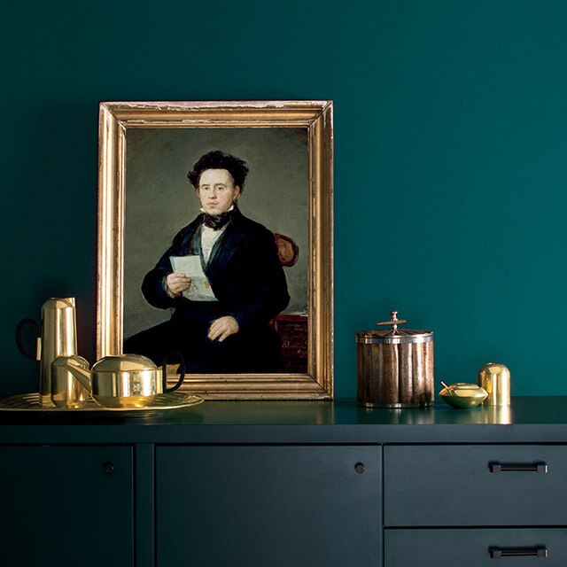 A dining room with green walls and a navy cabinet, featuring a portrait in a gilded frame and gold tea set.