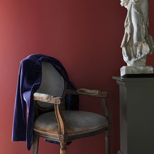 Pared pintada de rojo detrás de una estatua blanca sobre un pedestal y una silla tradicional de madera con un blazer púrpura en el respaldo.