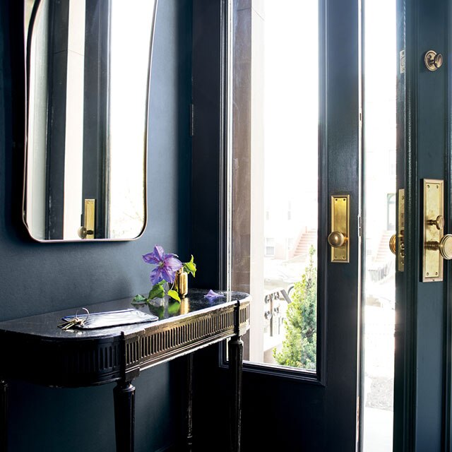 A dark blue entryway with mirror, door with large windows, and an accent table with flowers on it.