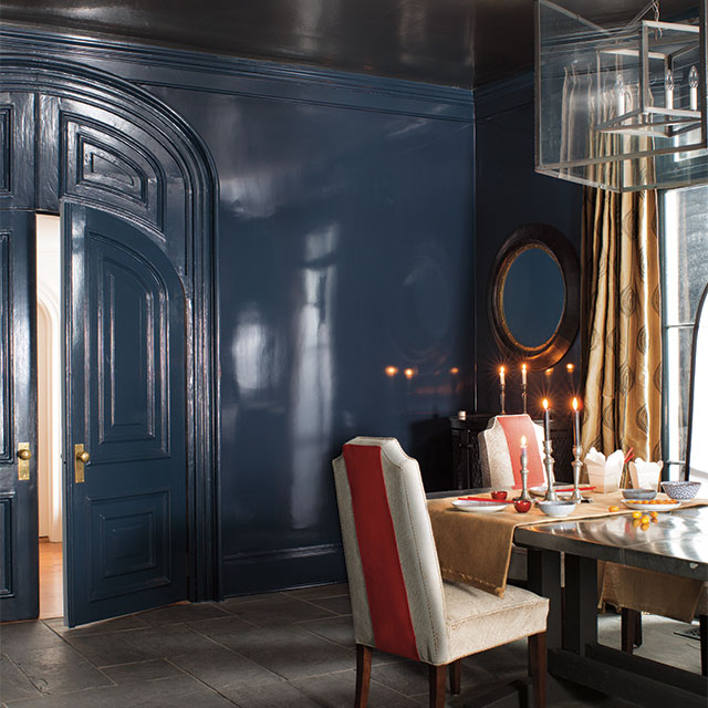 Dining room with glossy blue walls with a dining room table set with candlesticks & upholstered chairs with orange accents.