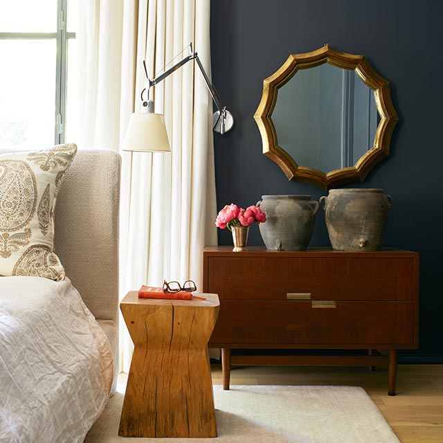 Una habitación pintada de negro con cama color crema, mesas decorativas de madera con macetas y flores, espejo dorado y cortinas blancas.