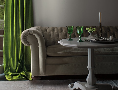 A living room with gray-painted walls, traditional gray couch, gray table with green accents, and green velvet drapes.