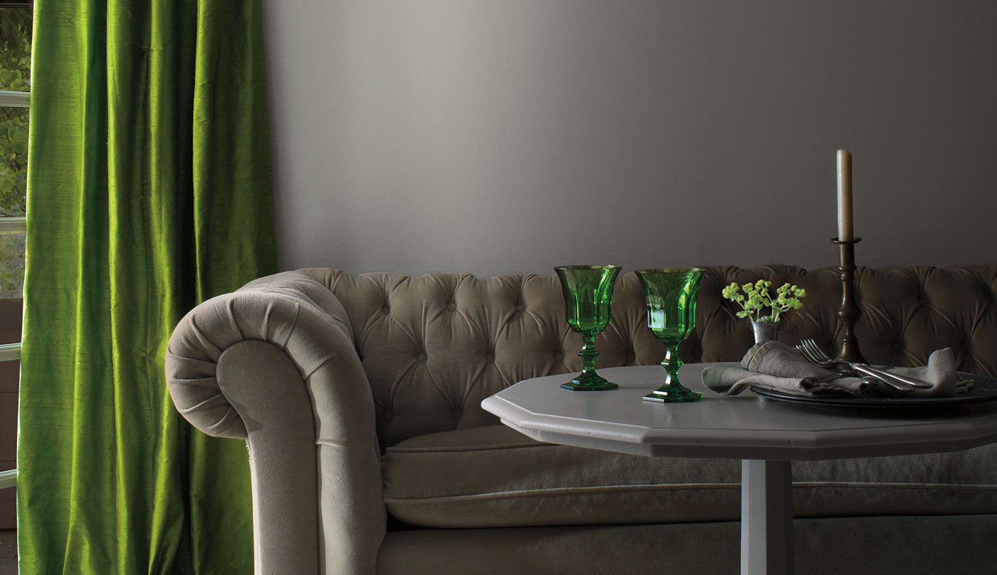 A living room with gray-painted walls, traditional gray couch, gray table with green accents, and green velvet drapes.