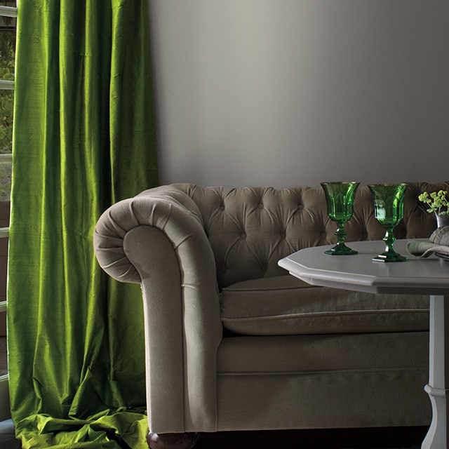A living room with gray-painted walls, traditional gray couch, gray table with green accents, and green velvet drapes.