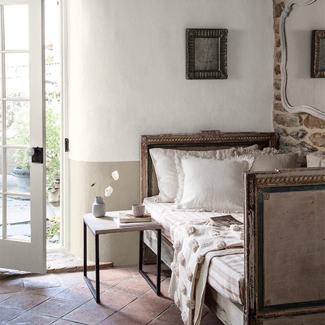 A French country bedroom with a white- and beige-painted wall, an open paned door, wood beamed ceiling, antique bed, and tiled floor.