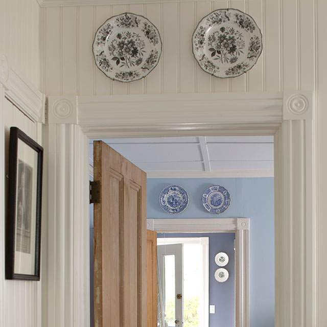 Une suite de couloirs peints en blanc, bleu clair et violet présentant des entrées de porte aux moulures blanches, des parquets en bois et la porte d’entrée de la maison tout au bout.