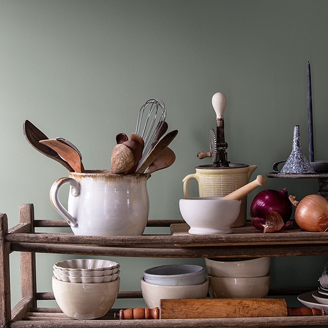 A collection of wooden utensils and other kitchenware on rustic shelves in front of a dark green-painted wall.