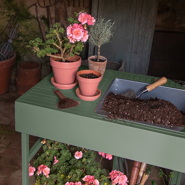 Un soporte para plantas de dos niveles pintado de verde con flores en macetas y herramientas de jardinería.