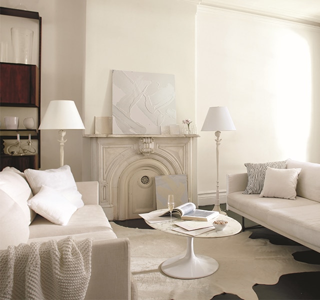 Salle de séjour neutre aux murs blancs avec une cheminée murale blanc cassé et une table basse ronde blanche entre deux canapés crème.