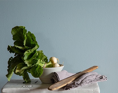 Verduras de hoja en un bol pequeño sobre una mesa con manteles y utensilios de madera frente a una pared pintada de azul.