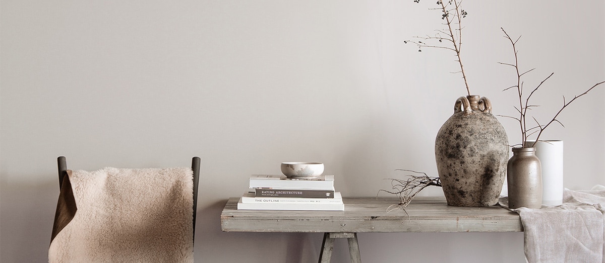 Salle à manger grise avec table rustique sur laquelle sont posés des livres, des vases et des branches, avec une chaise recouverte d’une couverture en fourrure blanche.