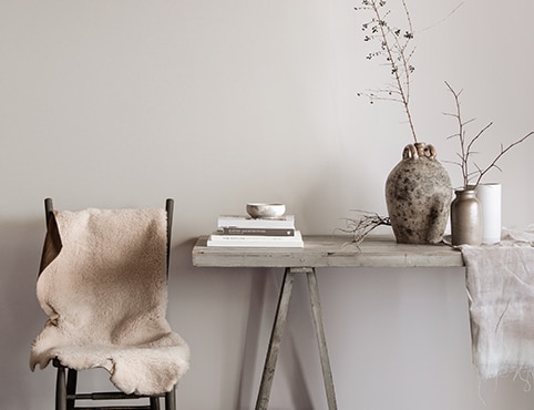 Salle à manger grise avec table rustique sur laquelle sont posés des livres, des vases et des branches, avec une chaise recouverte d’une couverture en fourrure blanche.