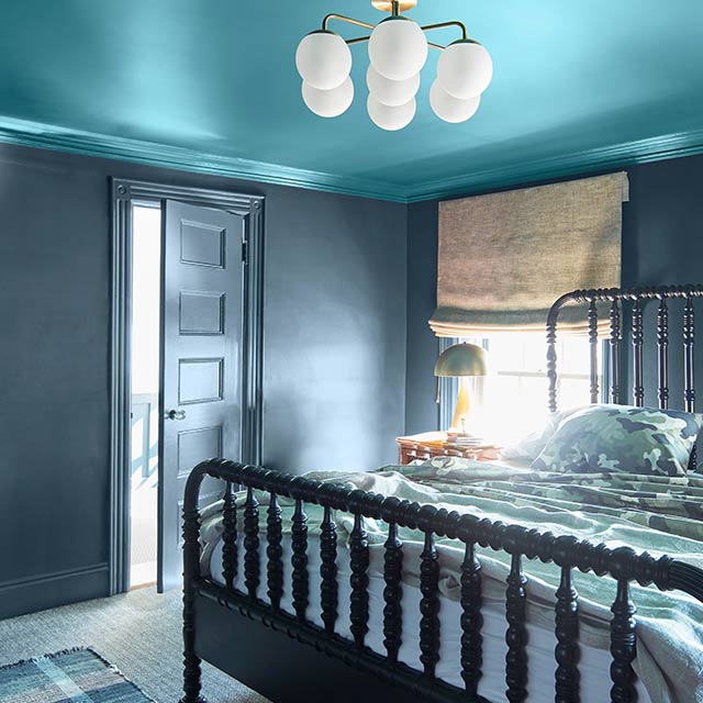 A bedroom with dark gray—blue painted walls, trim and door, a bright blue ceiling, and a black spindle bed.