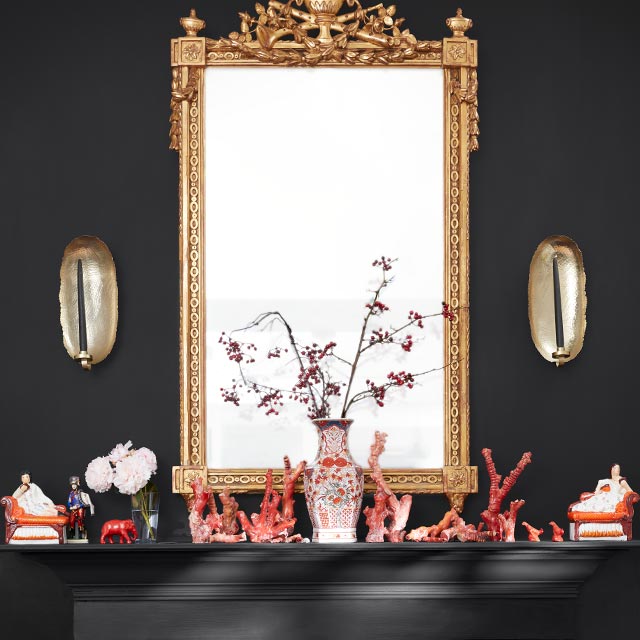 A black-painted wall and mantel with a multi-red pebble fireplace, a gold mirror and sconces, and red and white decorative items on the mantel.
