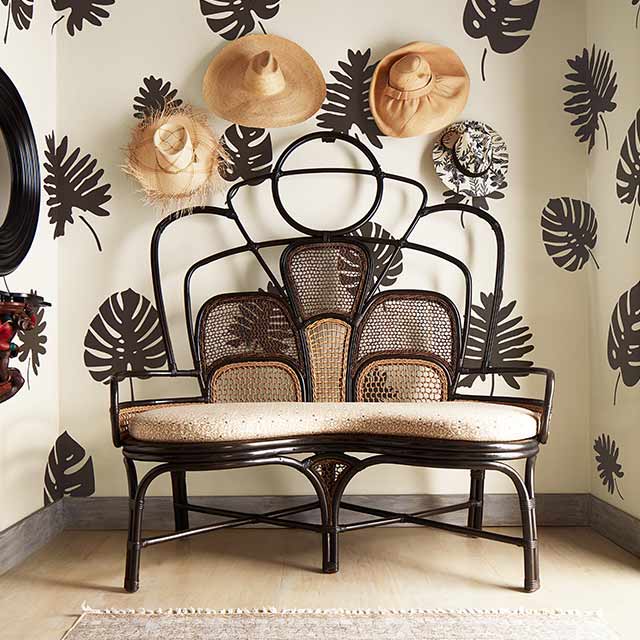 White painted walls with dark brown palm leaf stencils and straw hats hanging above a rattan and cloth loveseat, and a beige rug.