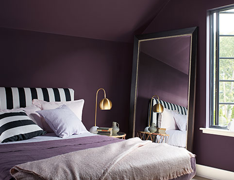 Chambre à coucher violet foncé avec moulures blanches, tête de lit et coussin à rayures noir et blanc, et couvertures mauves et blanches.