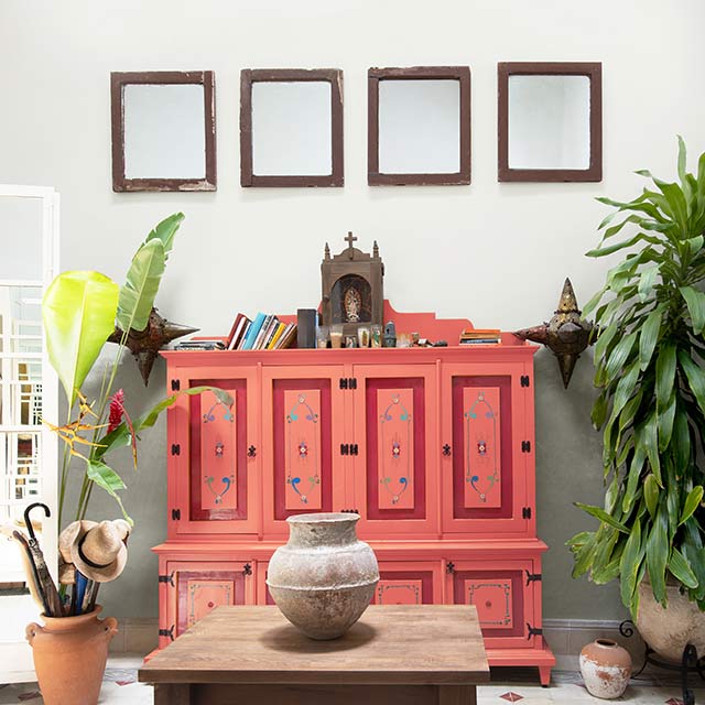 A coral pink-painted dresser in front of a white wall with framed mirrors overhead and house plants.